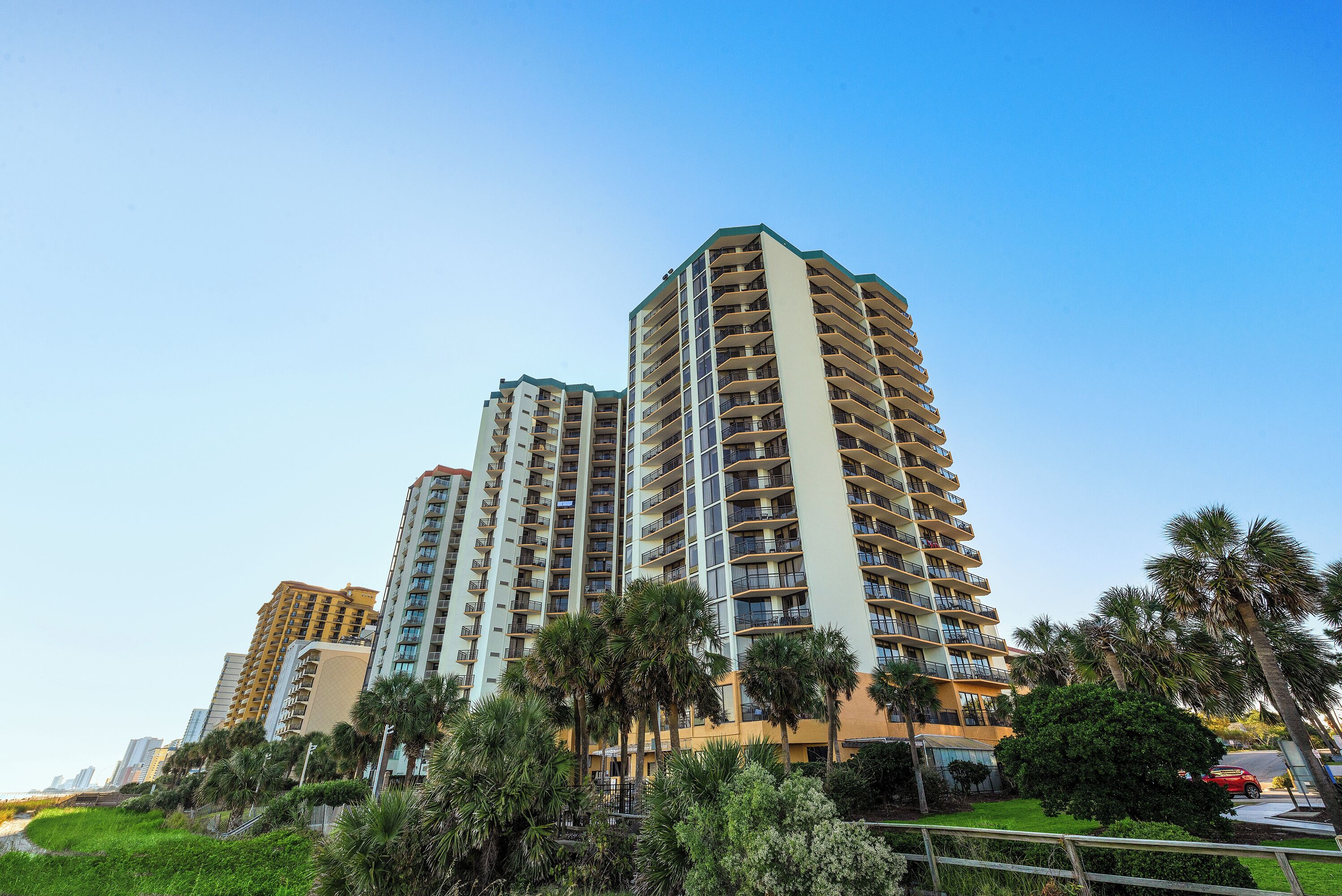 A high-rise residential building with multiple balconies, surrounded by palm trees and greenery.