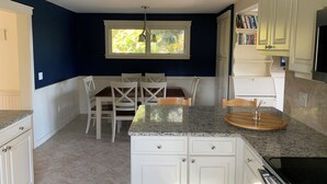 A kitchen with white cabinets, a granite countertop, a dining area with white chairs, and a window with a view of greenery. - Oceanfront Cape with dock on Bass Harbor (Bernard)
