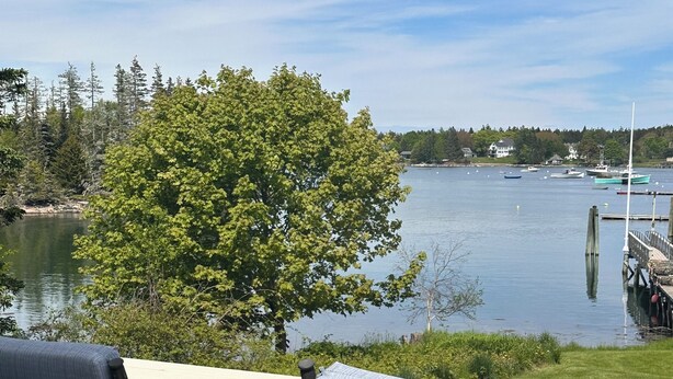A deck with two chairs facing a lake, surrounded by trees and a dock with boats. - Oceanfront Cape with dock on Bass Harbor (Bernard)
