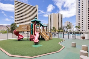Ein Spielplatz mit Rutschen und Klettergerüsten, umgeben von einem eingezäunten Bereich mit einem Schwimmbecken und hohen Gebäuden im Hintergrund.