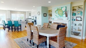 A modern kitchen with a central island, bar seating, and a dining area with wicker chairs. - Spacious, Luxe Design, Large Balcony & Incredible Views! (Tybee Island)