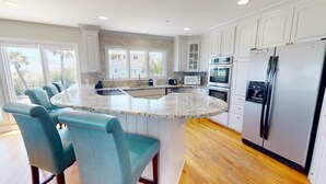 A modern kitchen with a central island, white cabinets, stainless steel appliances, and teal chairs. - Spacious, Luxe Design, Large Balcony & Incredible Views! (Tybee Island)