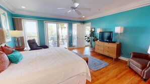 A bedroom with a large bed, a TV, a dresser, and a chair. There is a balcony with a view of palm trees. - Spacious, Luxe Design, Large Balcony & Incredible Views! (Tybee Island)