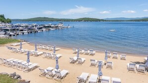 A beach with rows of sun loungers and umbrellas, a marina with boats, and a calm lake with a small boat in the distance. - 🏖 Lake Winnipesaukee Vacation at Akwa Marina 🏖 Beach, Pool, Hot Tub, Beach Bar (Laconia)
