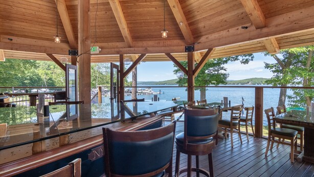 A wooden deck with a glass barrier overlooking a lake, featuring outdoor seating and a bar area. - 🏖 Lake Winnipesaukee Vacation at Akwa Marina 🏖 Beach, Pool, Hot Tub, Beach Bar (Laconia)
