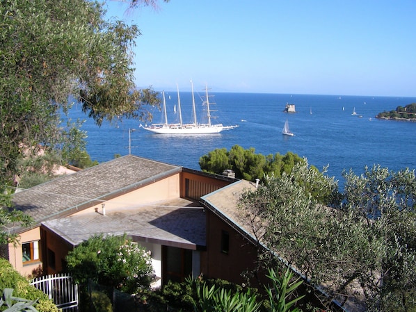 A coastal view with a large sailboat anchored in the distance, a building with a sloped roof, and dense greenery in the foreground.