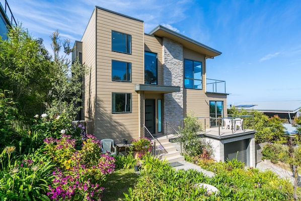 Une maison moderne à deux étages, dotée d’un balcon et entourée d’un jardin où poussent différentes plantes et fleurs.