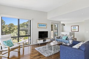 A modern living room with a large window, a flat-screen TV, a sofa, a coffee table, and a chair. - Bluview - Metres From The Beach, Views Included (Lorne)