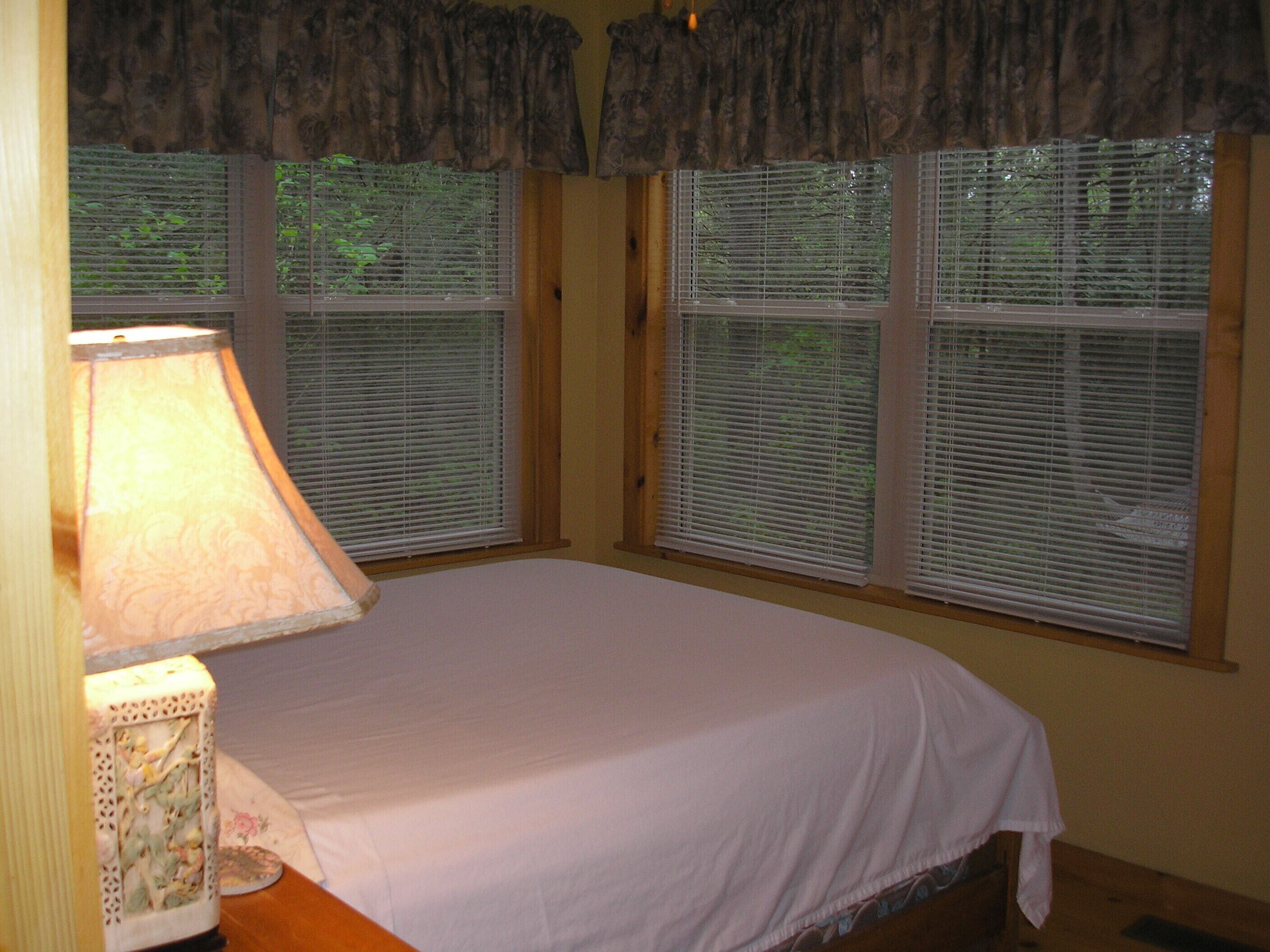 A bedroom with a bed, a lamp, and three windows with blinds.