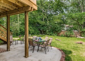 Eine überdachte Terrasse mit Betonboden und Holzsäulen, umgeben von einem üppigen Rasen und Bäumen.