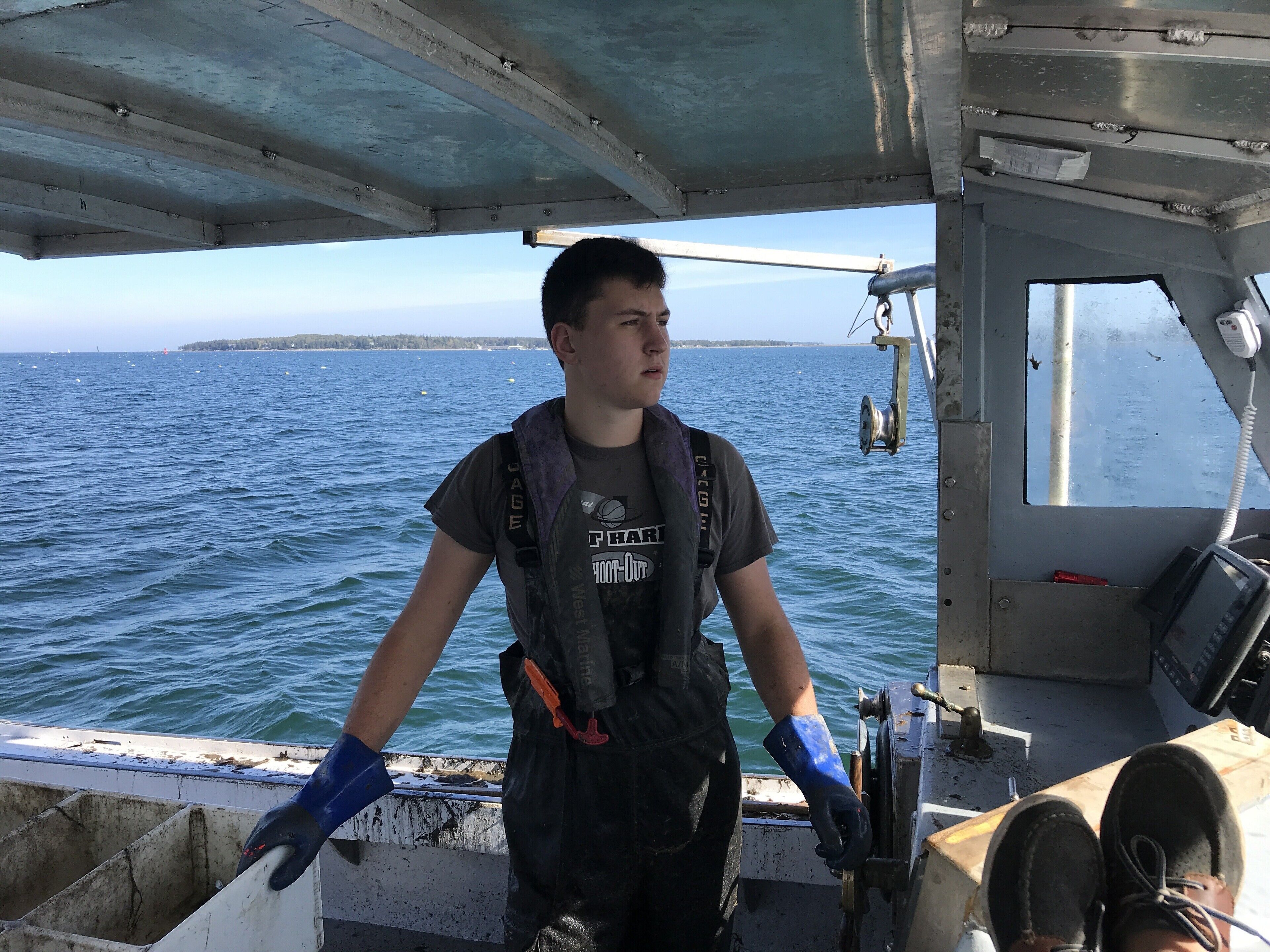 Una persona en un bote, con chaleco salvavidas y guantes, mirando el agua y una costa lejana.