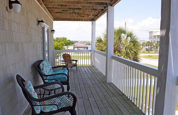 Une véranda couverte, avec deux fauteuils en osier et un banc, donnant sur un quartier résidentiel avec des maisons et un plan d’eau.