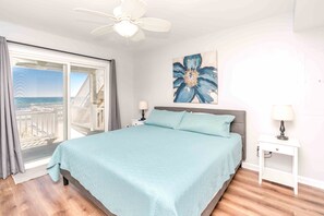 A bedroom with a bed, a nightstand, a ceiling fan, a painting, and a view of the ocean.