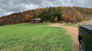 A rural setting with a house on a hill, surrounded by a grassy field and autumn-colored trees.