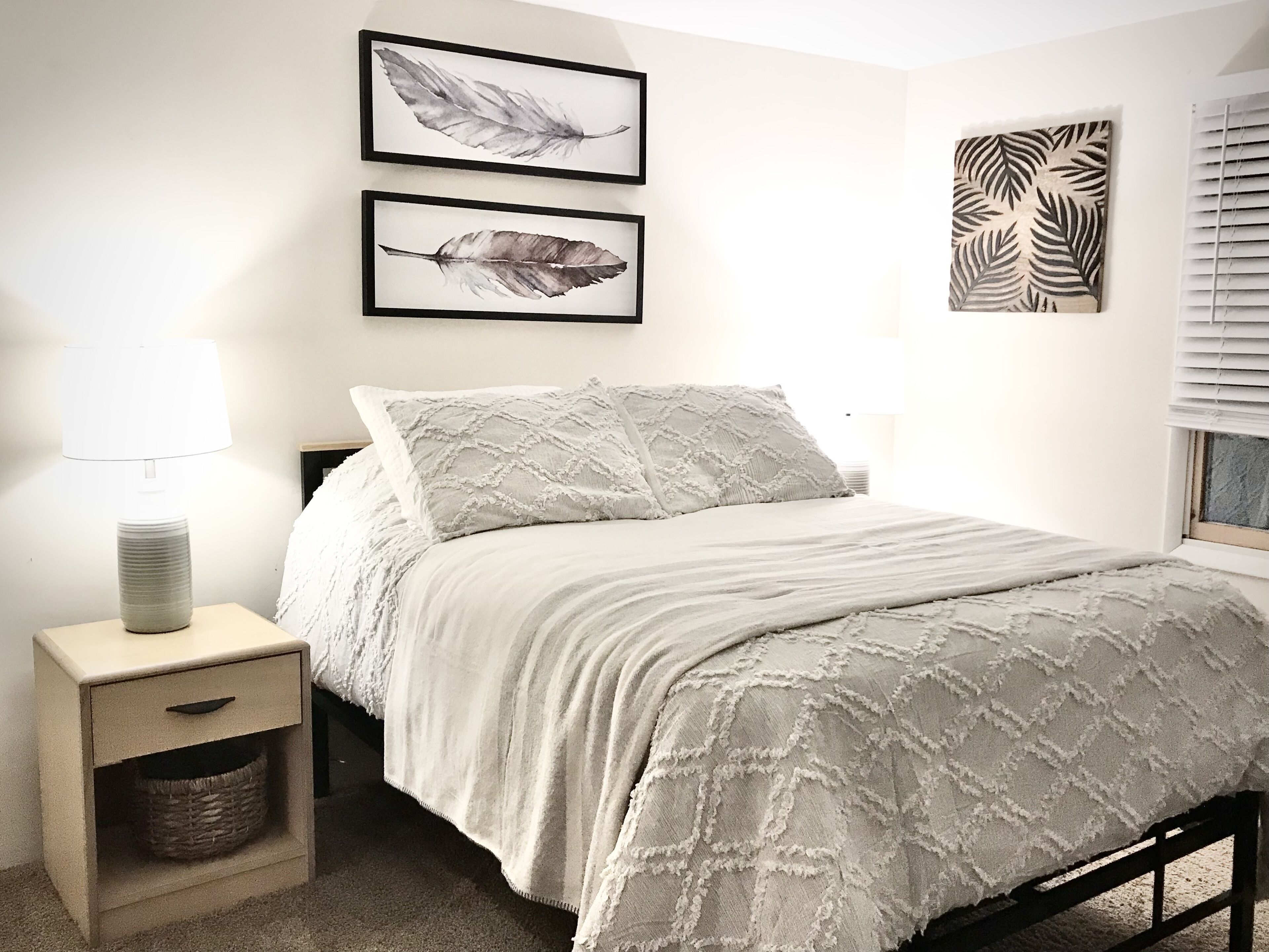 Un dormitorio con una cama, mesita de noche, lámpara y dos cuadros enmarcados en la pared.