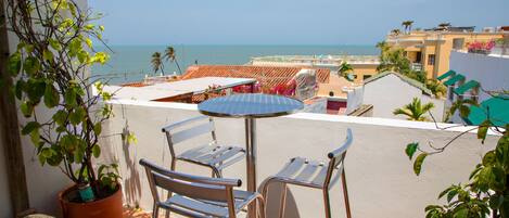 A balcony with a table and chairs, overlooking a coastal area with buildings and the sea.