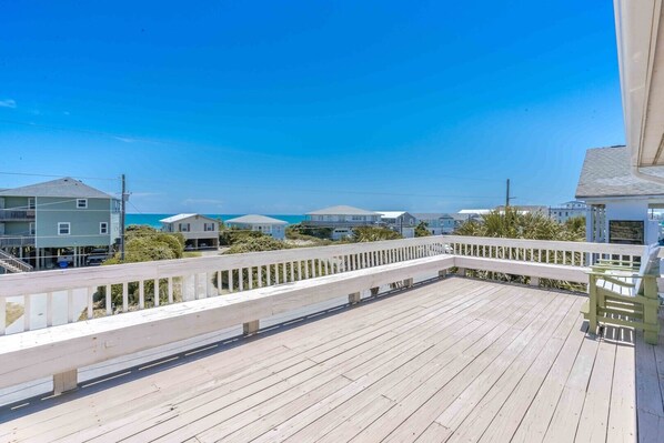 Una terraza de madera con barandal blanco, con vista a una playa con casas y el océano.