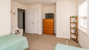 A bedroom with two beds, a dresser, a television, and a shelf with decorative items. - Flip Flops - Cozy 2nd Row Home - Ocean Views! (Surf City)