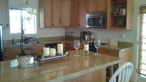 A kitchen with wooden cabinets, a stainless steel microwave, a marble countertop, and a dining area with white chairs. - MAGICAL 5-STAR RATED OCEAN VIEW CUSTOM HOME - PLEASE READ ALL 400 GUEST REVIEWS! (Summerland)