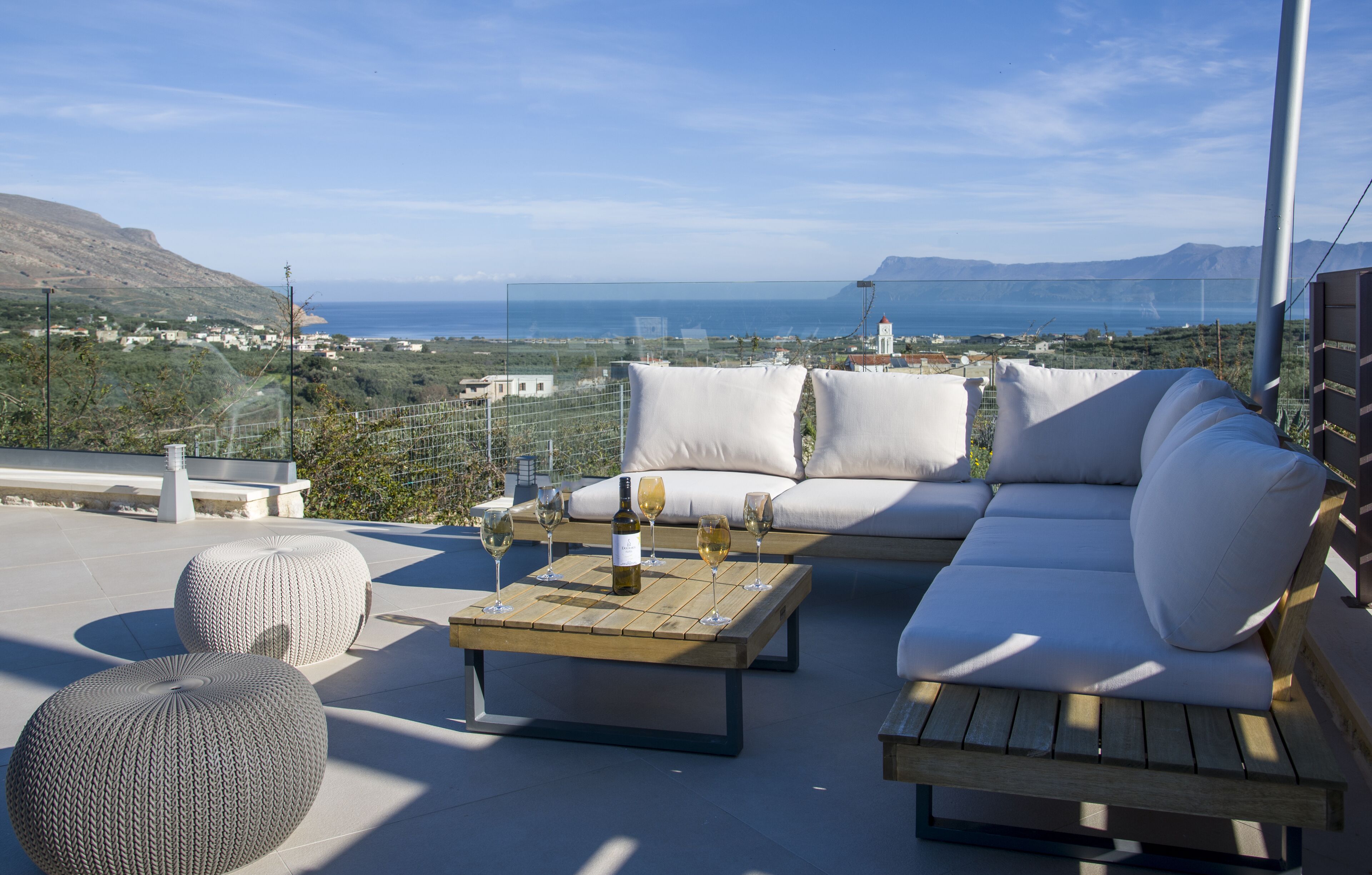 Una terraza con sofá, otomanas y una mesa con copas de vino y una botella.