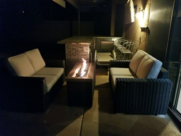 A cozy outdoor seating area with a fire feature, wicker furniture, and a brick accent wall. - Beautiful Modern Home Fully Upgraded! (Mesa)