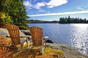 Two Adirondack chairs facing a lake with a forested shoreline.