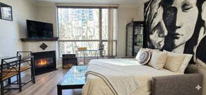 Un dormitorio con cama, televisión, chimenea, ventana grande y un cuadro.