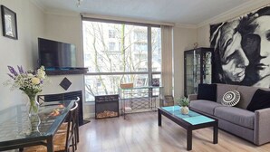 Een woonkamer met een glazen salontafel, een grijze bank, een televisie en een groot schilderij aan de muur.