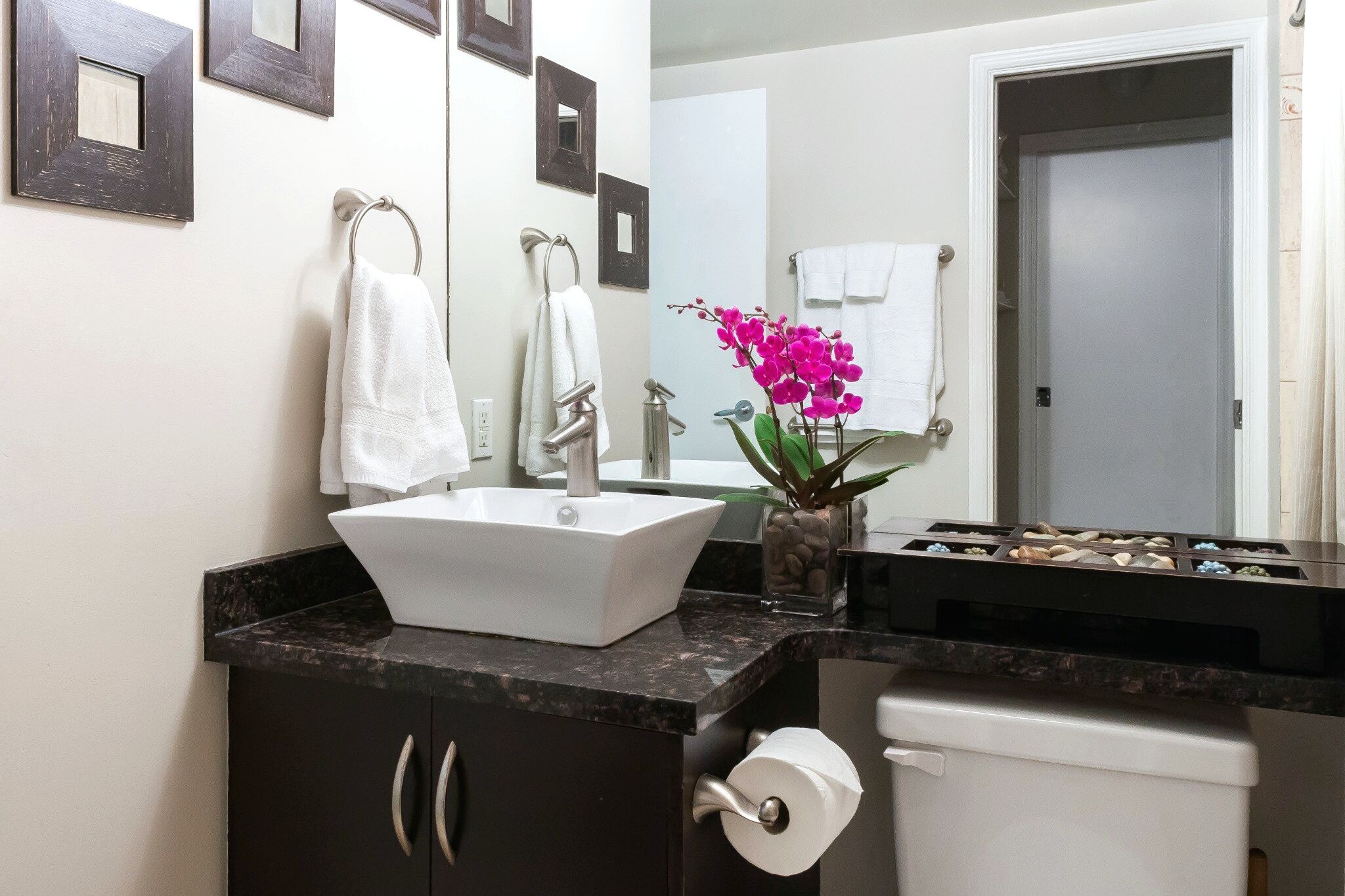 A bathroom with a white sink, dark cabinets, a toilet, and a mirror with framed pictures.