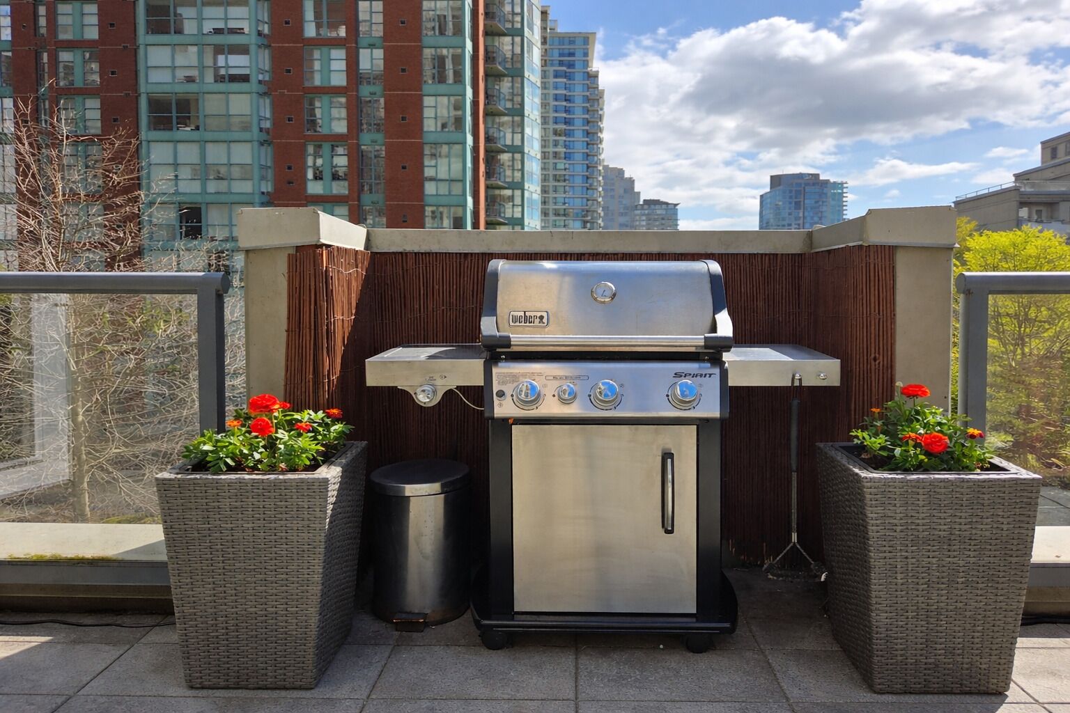 Un barbecue Weber Spirit portable, doté d’une finition en acier inoxydable, est placé sur un balcon, à côté de jardinières en osier contenant des fleurs rouges.