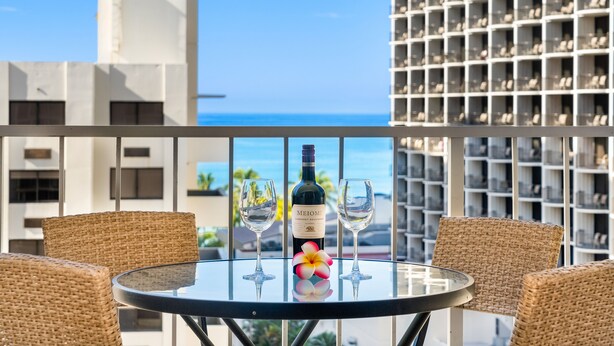Een balkon met een ronde tafel, twee rieten stoelen, een fles Meomi wijn, twee wijnglazen en een bloem.