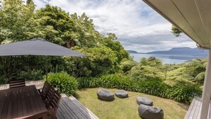 Una terraza de madera con mesa y sillas, rodeada de abundante vegetación y vista al mar.