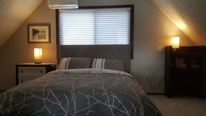 Et soveværelse med seng, natbord, lampe, bogreol og et vindue med skodder.