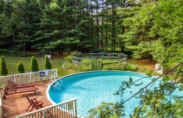 A swimming pool surrounded by a deck with lounge chairs, a fenced deck area, and a wooded backdrop.