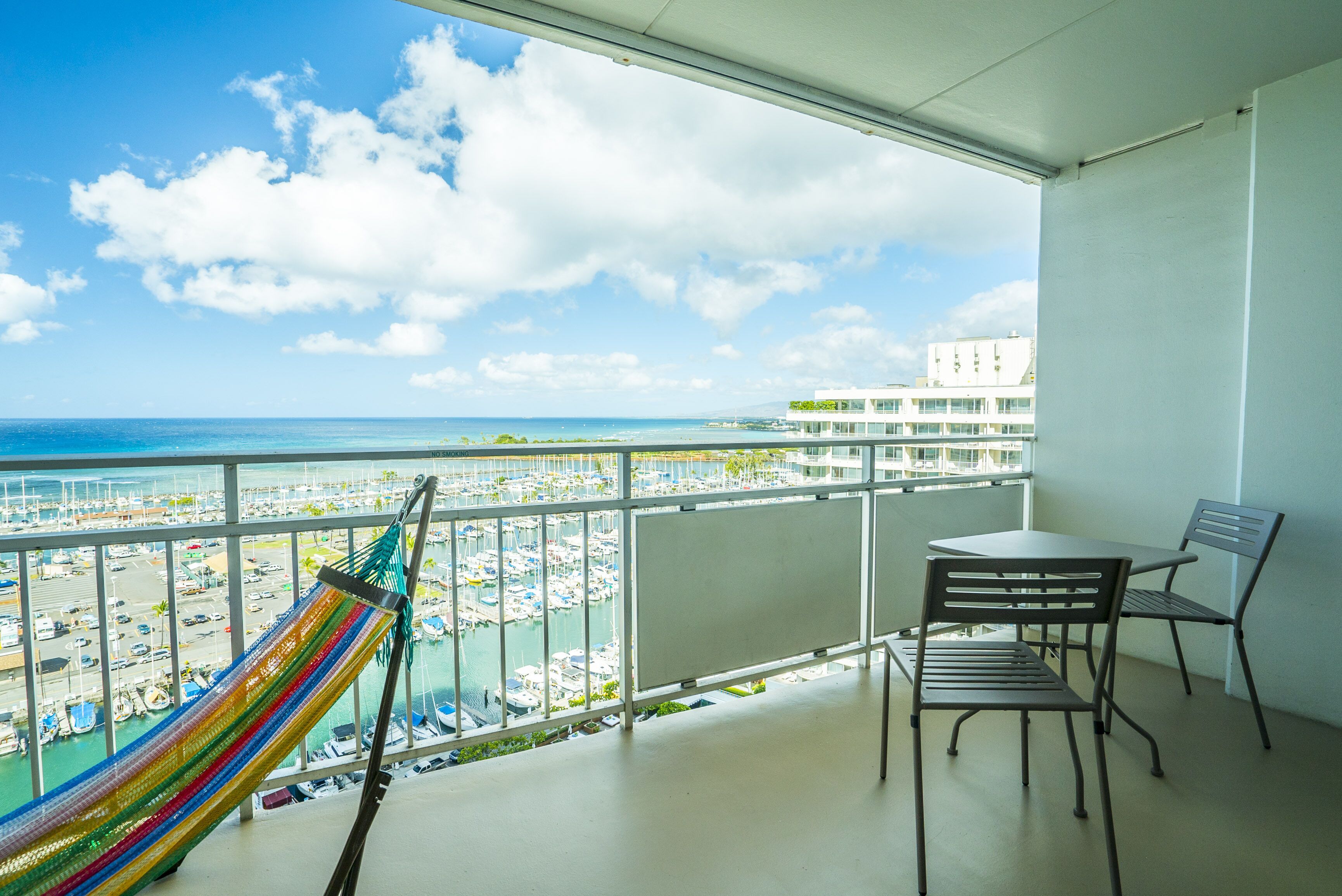 Un balcón con vista a un puerto deportivo y al océano, una hamaca y una mesa con sillas.