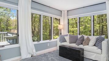 Un salon chaleureux avec un canapé gris, une commode noire et de grandes fenêtres donnant sur une forêt.