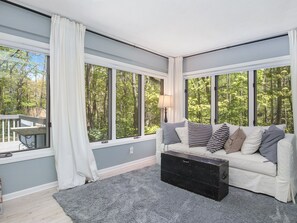 A cozy living room with a grey sofa, a black chest of drawers, and large windows offering a view of a forest.