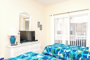 A hotel room with a television on a dresser, a bed with a blue patterned bedspread, a window with a view of a balcony, and a white wicker dresser. - Hoos at the Beach - Lovely 4 BR Sea Star Village Townhouse; Ocean View! (Surf City)