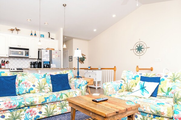 A living room with a sofa set, a coffee table, and a kitchen area with appliances. - Hoos at the Beach - Lovely 4 BR Sea Star Village Townhouse; Ocean View! (Surf City)