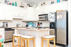 Une cuisine moderne avec des armoires blanches, un réfrigérateur en acier inoxydable, un four à micro-ondes et une machine à café. On y trouve également trois tabourets de bar et un mixeur sur le comptoir.