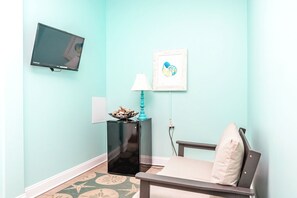 Une pièce avec un mur de couleur turquoise, un téléviseur à écran plat fixé au-dessus d’un meuble noir, une œuvre d’art encadrée, une lampe turquoise et une chaise blanche avec un coussin.
