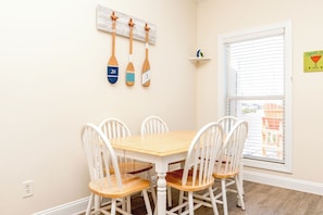 A dining area with a wooden table and white chairs, a wall-mounted number board, and a window with blinds. - Hoos at the Beach - Lovely 4 BR Sea Star Village Townhouse; Ocean View! (Surf City)