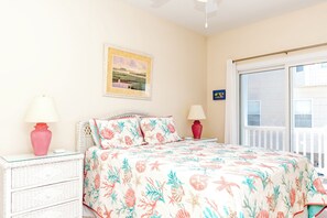 A bedroom with a bed, two bedside lamps, a wicker nightstand, a framed picture on the wall, and a window with a view. - Hoos at the Beach - Lovely 4 BR Sea Star Village Townhouse; Ocean View! (Surf City)
