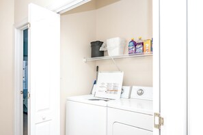 Un espace buanderie comprenant un lave-linge, une étagère avec des produits de nettoyage et une porte donnant sur une autre pièce.