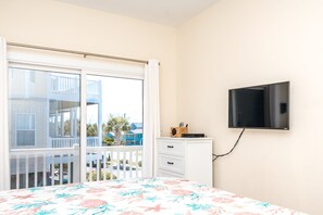 A bedroom with a bed, a TV mounted on the wall, a white dresser, and a balcony with a view of other buildings. - Hoos at the Beach - Lovely 4 BR Sea Star Village Townhouse; Ocean View! (Surf City)