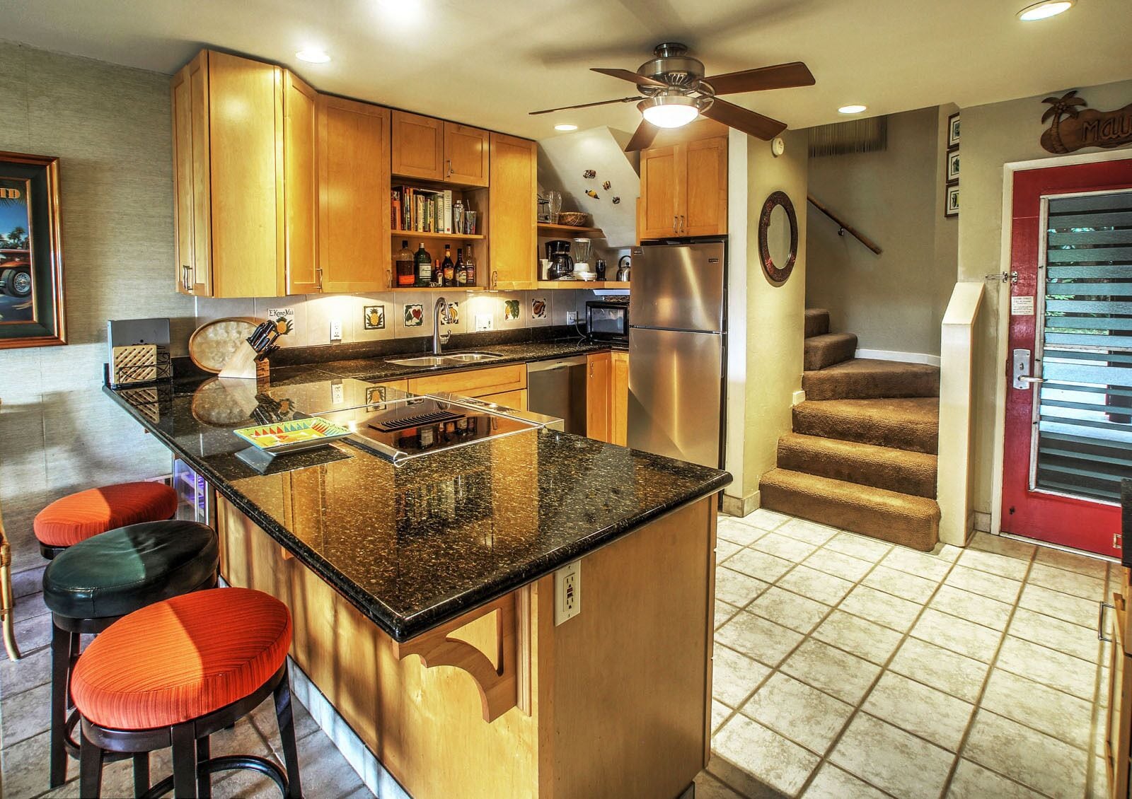 A kitchen with wooden cabinets, a stainless steel refrigerator, a microwave, and a granite countertop. There are two stools with red cushions.