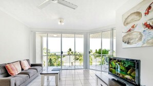 A spacious living room with a grey sofa, a coffee table, a flat-screen TV, and a balcony with glass doors. - Luxury Beach Front Accommodation  (Trinity Beach)