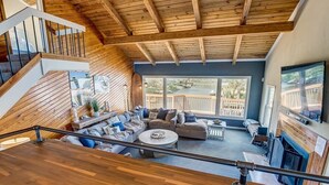 Een ruime woonkamer met een hoge plafond en houten balken, een groot raam met uitzicht op een terras, en een open haard.
