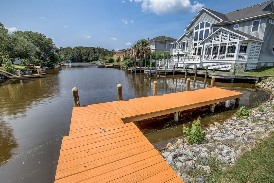 Un muelle de madera se adentra en un río tranquilo, rodeado de abundante vegetación y edificios residenciales.