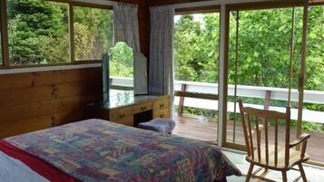 Une chambre à coucher comprenant un lit, une commode, un téléviseur et offrant une vue sur la verdure grâce à de grandes fenêtres.